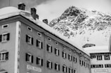 A winter scene with a historic hotel building covered in snow, set against a backdrop of a rugged, snow-capped mountain. The architecture features multiple windows with dark shutters and decorative moldings. A sign reads 'HOTEL RAVIZZA' on the side of the building.