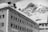 A winter scene with a historic hotel building covered in snow, set against a backdrop of a rugged, snow-capped mountain. The architecture features multiple windows with dark shutters and decorative moldings. A sign reads 'HOTEL RAVIZZA' on the side of the building.