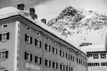 A winter scene with a historic hotel building covered in snow, set against a backdrop of a rugged, snow-capped mountain. The architecture features multiple windows with dark shutters and decorative moldings. A sign reads 'HOTEL RAVIZZA' on the side of the building.