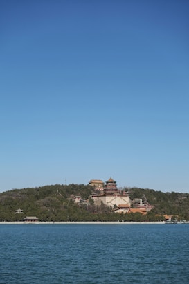 A serene landscape features a traditional Chinese palace complex situated on a lush, green hill overlooking a calm body of water. The architecture includes ornate structures with multilayered roofs and intricate detailing. The sky is clear and blue, enhancing the peaceful ambiance.