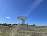 A large radio telescope stands on a grassy hill under a clear blue sky. The structure consists of a massive dish supported by a metal framework, surrounded by open fields and scattered trees.