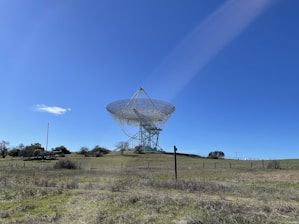 A large radio telescope stands on a grassy hill under a clear blue sky. The structure consists of a massive dish supported by a metal framework, surrounded by open fields and scattered trees.