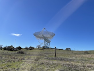 A large radio telescope stands on a grassy hill under a clear blue sky. The structure consists of a massive dish supported by a metal framework, surrounded by open fields and scattered trees.