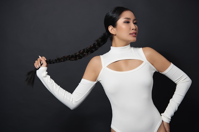 A woman with long, braided hair wearing a white cut-out top poses against a dark background. She holds the end of her braid in one hand while striking a confident pose. The attire features open shoulder sleeves and a high neckline.