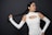 A woman with long, braided hair wearing a white cut-out top poses against a dark background. She holds the end of her braid in one hand while striking a confident pose. The attire features open shoulder sleeves and a high neckline.