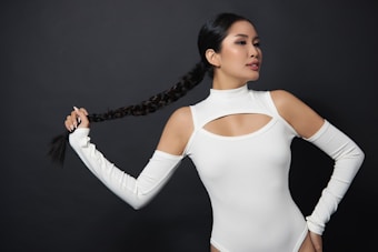 A woman with long, braided hair wearing a white cut-out top poses against a dark background. She holds the end of her braid in one hand while striking a confident pose. The attire features open shoulder sleeves and a high neckline.