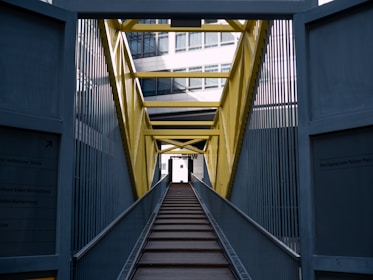An industrial bridge with a bright yellow framework and metal sides leads into the distance. The pathway is made of wooden planks and is flanked by tall metal barriers. The architecture of the bridge contrasts with modern glass buildings partially visible in the background.