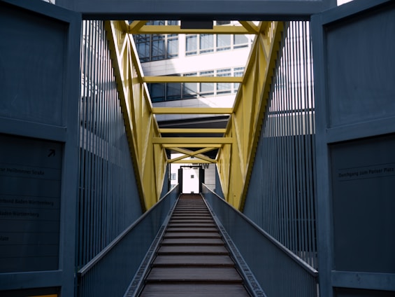 An industrial bridge with a bright yellow framework and metal sides leads into the distance. The pathway is made of wooden planks and is flanked by tall metal barriers. The architecture of the bridge contrasts with modern glass buildings partially visible in the background.