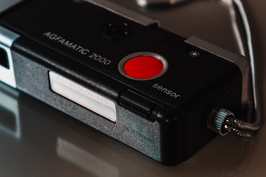 A close-up shot of a vintage AGFAMATIC 2000 camera. The image focuses on the details of the camera, highlighting features such as the large red button labeled 'sensor' and the silver connector cable. A white label on the side reads 'AGFACOLOR 110'. The camera casing is predominantly black with a sleek design.