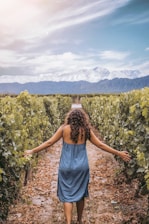 A woman in a blue dress strides through a lush vineyard with grape vines on both sides. Her arms are outstretched, gently touching the leaves. In the background, snow-capped mountains rise under a bright, slightly cloudy sky.