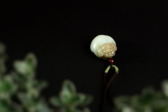A white and beige carved pendant with a round shape featuring a small figure, likely made of jade or similar material, is seen against a dark background. A red bead is attached to the bottom with green and brown strings. The foreground is blurred with green leaves out of focus.