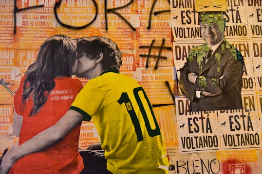 A street art mural featuring a young couple kissing, the girl wearing a red shirt and the boy in a yellow soccer jersey with the number 10. The background consists of protest posters with bold text and the image of a smiling man in a suit with green slime dripping over him. The art is layered on a wall covered in text and graffiti.
