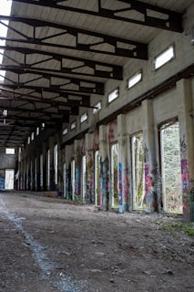 An abandoned building with a high ceiling supported by exposed wooden beams. The walls are adorned with colorful graffiti art. The floor is covered in dirt and rubble, leading to an open doorway at the far end. Natural light filters in through upper windows, creating a partially illuminated corridor.