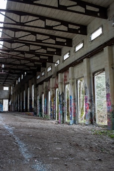 An abandoned building with a high ceiling supported by exposed wooden beams. The walls are adorned with colorful graffiti art. The floor is covered in dirt and rubble, leading to an open doorway at the far end. Natural light filters in through upper windows, creating a partially illuminated corridor.