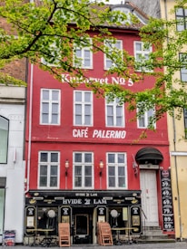 A red and white multi-story building houses 'Café Palermo,' with large rectangular windows and a ground-floor entrance. The facade is adorned with text and small lantern-like lights. Chairs are stacked near the entrance, suggesting an outdoor seating area. Above, green leaves from nearby trees frame the building.