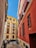 A narrow alleyway lined with vibrant buildings, one painted in bright red and another in yellow with white trim. Numerous balconies with wrought iron railings overlook the street. Several motorcycles are parked along the side, and a Spanish flag hangs in one of the windows. The sky is clear and blue above.