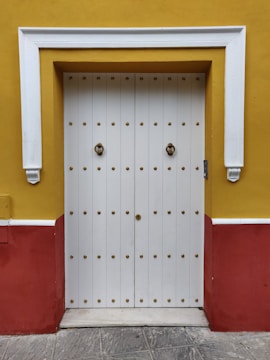A traditional wooden door with ornate brass studs set in a vibrant yellow wall, accented by a red base and decorative white trim framing the door.