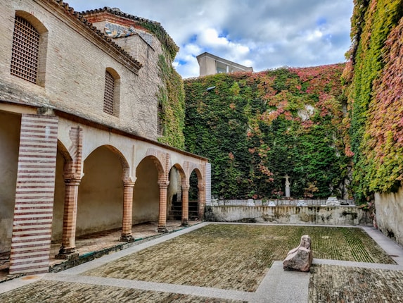 A serene courtyard surrounded by tall walls covered in lush green and red ivy. The space features an arched colonnade with brick and stone architecture on one side, leading to an open area with a stone pathway and a small statue visible against the back wall.