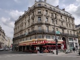 A classic Parisian-style building with intricate architectural details, including ornate balconies and arched windows. The ground floor houses a brasserie with a red and yellow striped awning and outdoor seating. Several people can be seen walking along the sidewalk, and a few vehicles are parked nearby.