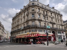 A classic Parisian-style building with intricate architectural details, including ornate balconies and arched windows. The ground floor houses a brasserie with a red and yellow striped awning and outdoor seating. Several people can be seen walking along the sidewalk, and a few vehicles are parked nearby.