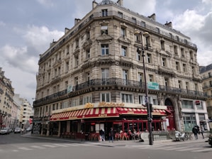 A classic Parisian-style building with intricate architectural details, including ornate balconies and arched windows. The ground floor houses a brasserie with a red and yellow striped awning and outdoor seating. Several people can be seen walking along the sidewalk, and a few vehicles are parked nearby.