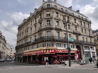 A classic Parisian-style building with intricate architectural details, including ornate balconies and arched windows. The ground floor houses a brasserie with a red and yellow striped awning and outdoor seating. Several people can be seen walking along the sidewalk, and a few vehicles are parked nearby.