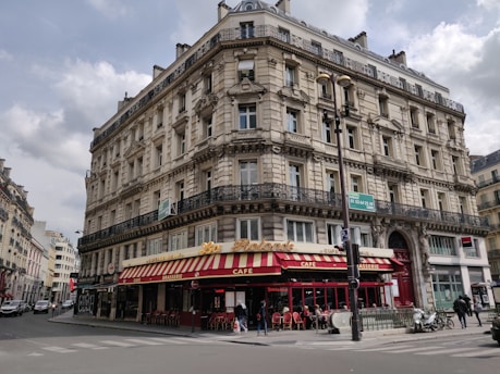 A classic Parisian-style building with intricate architectural details, including ornate balconies and arched windows. The ground floor houses a brasserie with a red and yellow striped awning and outdoor seating. Several people can be seen walking along the sidewalk, and a few vehicles are parked nearby.