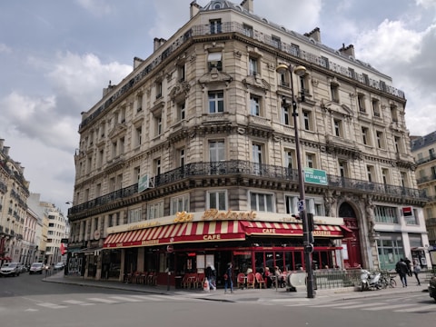 A classic Parisian-style building with intricate architectural details, including ornate balconies and arched windows. The ground floor houses a brasserie with a red and yellow striped awning and outdoor seating. Several people can be seen walking along the sidewalk, and a few vehicles are parked nearby.