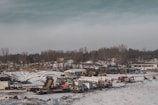 A snowy landscape features a construction or industrial site with several pieces of heavy machinery and vehicles such as trucks and excavators. The ground is partially covered with snow, and there's a backdrop of barren trees. The sky is overcast, providing a muted, cold atmosphere.