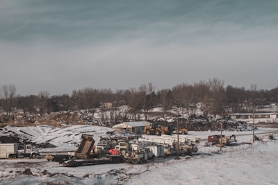 A snowy landscape features a construction or industrial site with several pieces of heavy machinery and vehicles such as trucks and excavators. The ground is partially covered with snow, and there's a backdrop of barren trees. The sky is overcast, providing a muted, cold atmosphere.