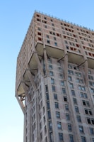 A tall, modernist building with a unique structural design characterized by visible supporting columns. The architecture features a mix of concrete and glass elements, with numerous rectangular windows arranged in a grid pattern. The building's facade exhibits a repetitive and geometric aesthetic, and the overall construction suggests a brutalist style.