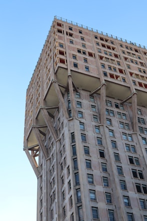 A tall, modernist building with a unique structural design characterized by visible supporting columns. The architecture features a mix of concrete and glass elements, with numerous rectangular windows arranged in a grid pattern. The building's facade exhibits a repetitive and geometric aesthetic, and the overall construction suggests a brutalist style.