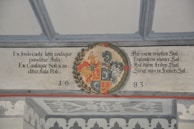 A detailed heraldic mural featuring a coat of arms situated in the center with intricate floral and scroll elements surrounding it. Latin and German text is inscribed on either side of the coat of arms, dated with the year 1683 beneath.