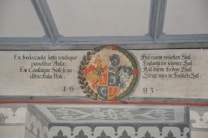 A detailed heraldic mural featuring a coat of arms situated in the center with intricate floral and scroll elements surrounding it. Latin and German text is inscribed on either side of the coat of arms, dated with the year 1683 beneath.