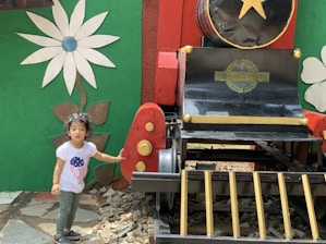 A child in a white t-shirt with a blue and red design stands next to a colorful, decorative train structure. The setting includes a large flower mural on a green wall, and the train is painted in red, black, and gold with a large yellow star on the front.