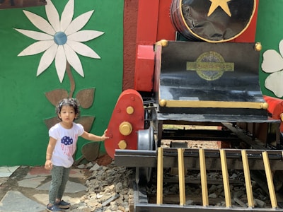A child in a white t-shirt with a blue and red design stands next to a colorful, decorative train structure. The setting includes a large flower mural on a green wall, and the train is painted in red, black, and gold with a large yellow star on the front.
