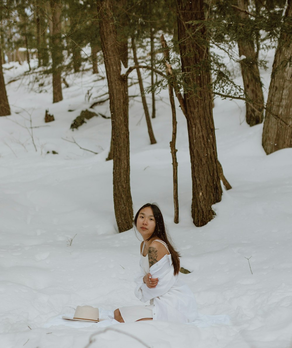 woman in white dress sitting on snow covered ground near brown bare trees during daytime