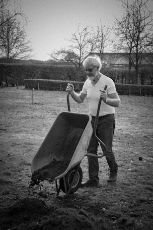 grayscale photo of man in long sleeve shirt and pants holding a wheel barrow