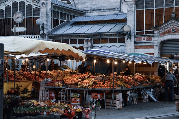A bustling outdoor market scene with a variety of fruits and vegetables displayed on tables under a striped canopy. The market is set against the backdrop of an ornate building with arched windows and decorative elements. Several people are browsing the stalls, contributing to a lively atmosphere.