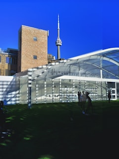 A tall communications tower rises against a clear blue sky, with a brick building and a large transparent tent-like structure in the foreground. Shadows of trees are visible on the ground, and several people are standing or walking on the grass.