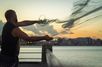 A fisherman casting a net into the sparkling blue sea at sunset.