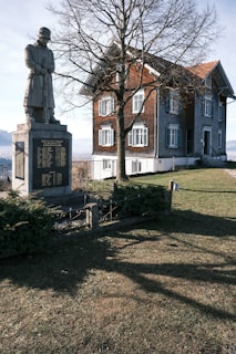 A stone statue of a soldier stands on a pedestal with engraved text in front of a tall, bare tree. Behind the tree is a traditional brick and stone house with green shutters, situated on a grassy landscape under a clear blue sky. Shadows are cast across the grass, indicating sunlight in the scene.