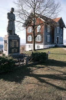 A stone statue of a soldier stands on a pedestal with engraved text in front of a tall, bare tree. Behind the tree is a traditional brick and stone house with green shutters, situated on a grassy landscape under a clear blue sky. Shadows are cast across the grass, indicating sunlight in the scene.