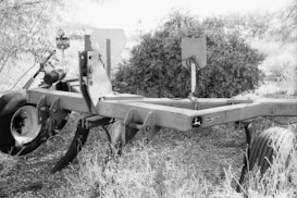 A grayscale image featuring a large piece of agricultural machinery with wheels. The equipment appears to be intended for soil management, as indicated by a logo and text. It is positioned outdoors, surrounded by dry vegetation and a bush in the background.