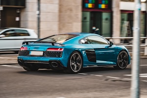 A sleek, turquoise sports car drives down a city street. The car has a modern design with black accents and a low profile, suggestive of high speed and performance. In the background, blurred pedestrians and urban buildings create a sense of motion and city life.