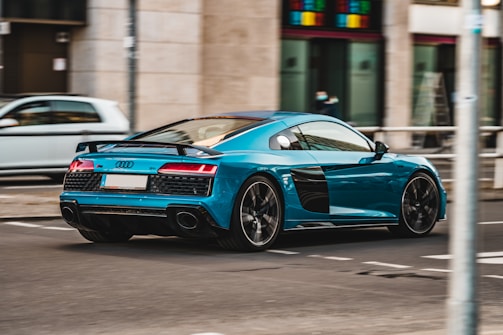A sleek, turquoise sports car drives down a city street. The car has a modern design with black accents and a low profile, suggestive of high speed and performance. In the background, blurred pedestrians and urban buildings create a sense of motion and city life.