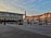 An urban plaza with a wide paved area is surrounded by modern buildings. The facade of a large building features historical images and a 'Bucovina 100' sign. The sky is partly cloudy and filled with scattered birds.