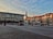 An urban plaza with a wide paved area is surrounded by modern buildings. The facade of a large building features historical images and a 'Bucovina 100' sign. The sky is partly cloudy and filled with scattered birds.