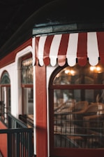A vintage-style train carriage featuring red and white striped awnings above its windows. The carriage is bordered by a blue railing, and warm lights illuminate the interior which appears to have wooden chairs. The ambiance is nostalgic and cozy, possibly evoking memories of past travels.