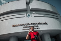 A person wearing sunglasses and a red shirt stands in front of a building with large, bold letters in Cyrillic on its facade. The building has a modern architectural style with curves and smooth surfaces. Bright sunlight casts shadows on the structure, and the sky is clear with a few clouds.